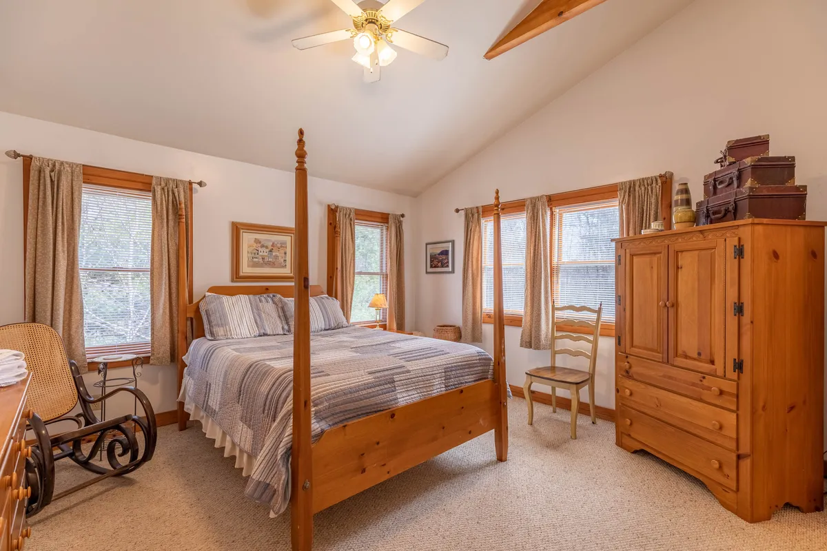 A welcoming bedroom with a classic four-poster bed, rocking chair, wooden dresser, and a window letting in soft natural light. The scene suggests peaceful mornings and leisurely moments spent in a tranquil, private retreat.