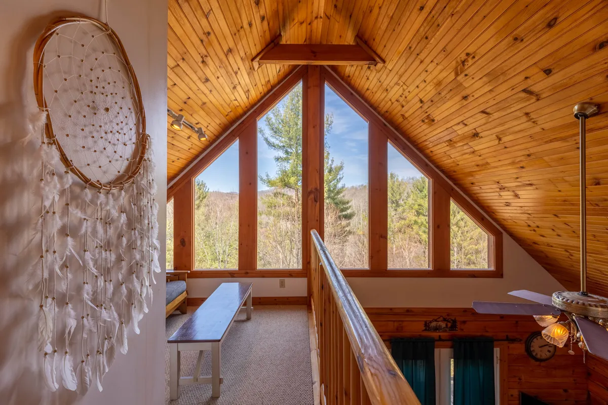 A peaceful seating area featuring a bench beneath large windows framed by wooden beams, letting in abundant natural light and offering serene views of the outdoors. A dreamcatcher hangs nearby, enhancing the rustic and nature-inspired atmosphere of the space. The scene feels open and connected to the natural surroundings.