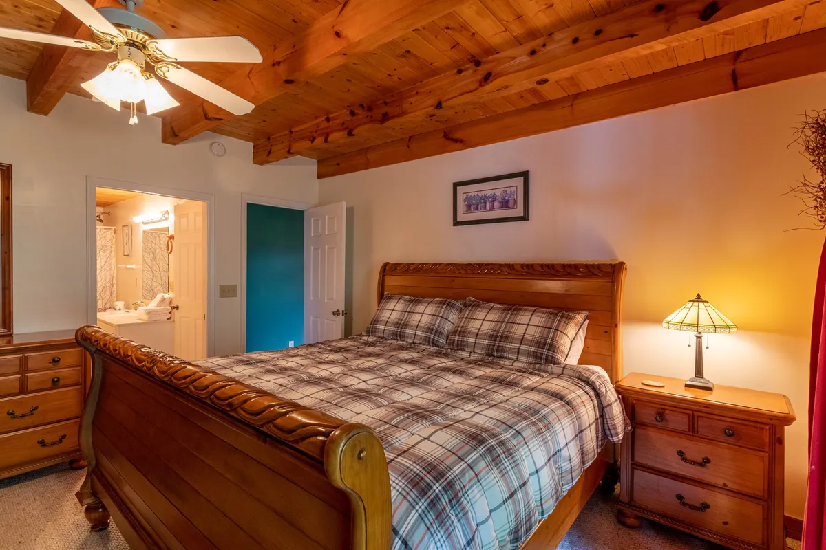 A warmly lit bedroom featuring a wooden bed frame adorned with plaid bedding, a matching nightstand with a lamp, a sturdy wooden dresser, and framed artwork on the wall. The inviting space radiates comfort and provides a sense of privacy and relaxation.