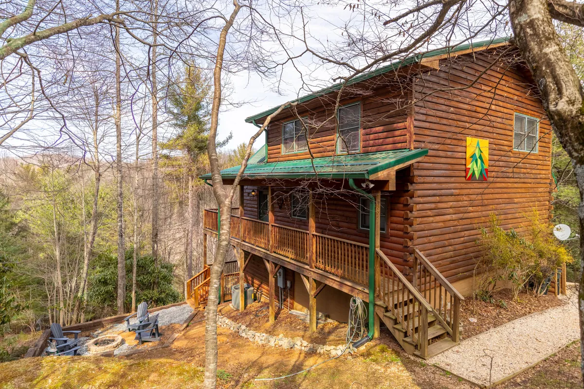 The hero image shows a charming log cabin nestled amidst lush trees, with a unique green roof, colorful wall art adding character, and an inviting deck perfect for sunset views. The cabin's exterior hints at welcoming spaces for outdoor gatherings, barbecues, and cozy evenings by the firepit, all set against the beautiful backdrop of Boone's rolling hills.