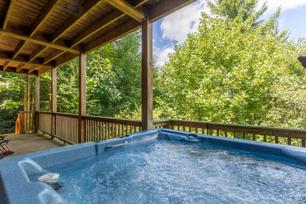 An inviting covered deck with a bubbling hot tub nestled amidst natural greenery. The wooden deck creates a transition between indoor fun and outdoor relaxation, with the tranquil setting perfect for unwinding after busy days.