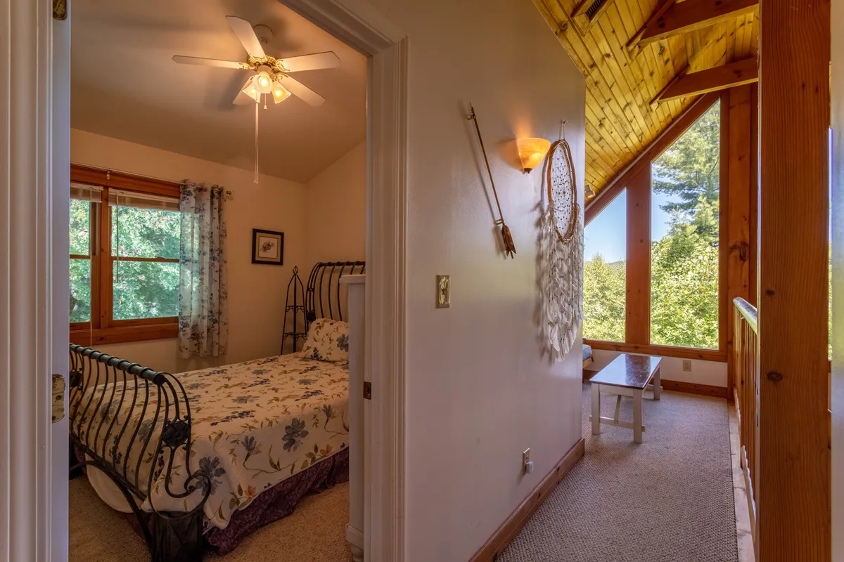 A tranquil bedroom with a floral bedspread, large windows that flood the space with natural light, and a wooden ceiling overhead. The view through the windows hints at the surrounding natural beauty and mountain landscape, while the calm, inviting decor invites relaxation and intimacy.