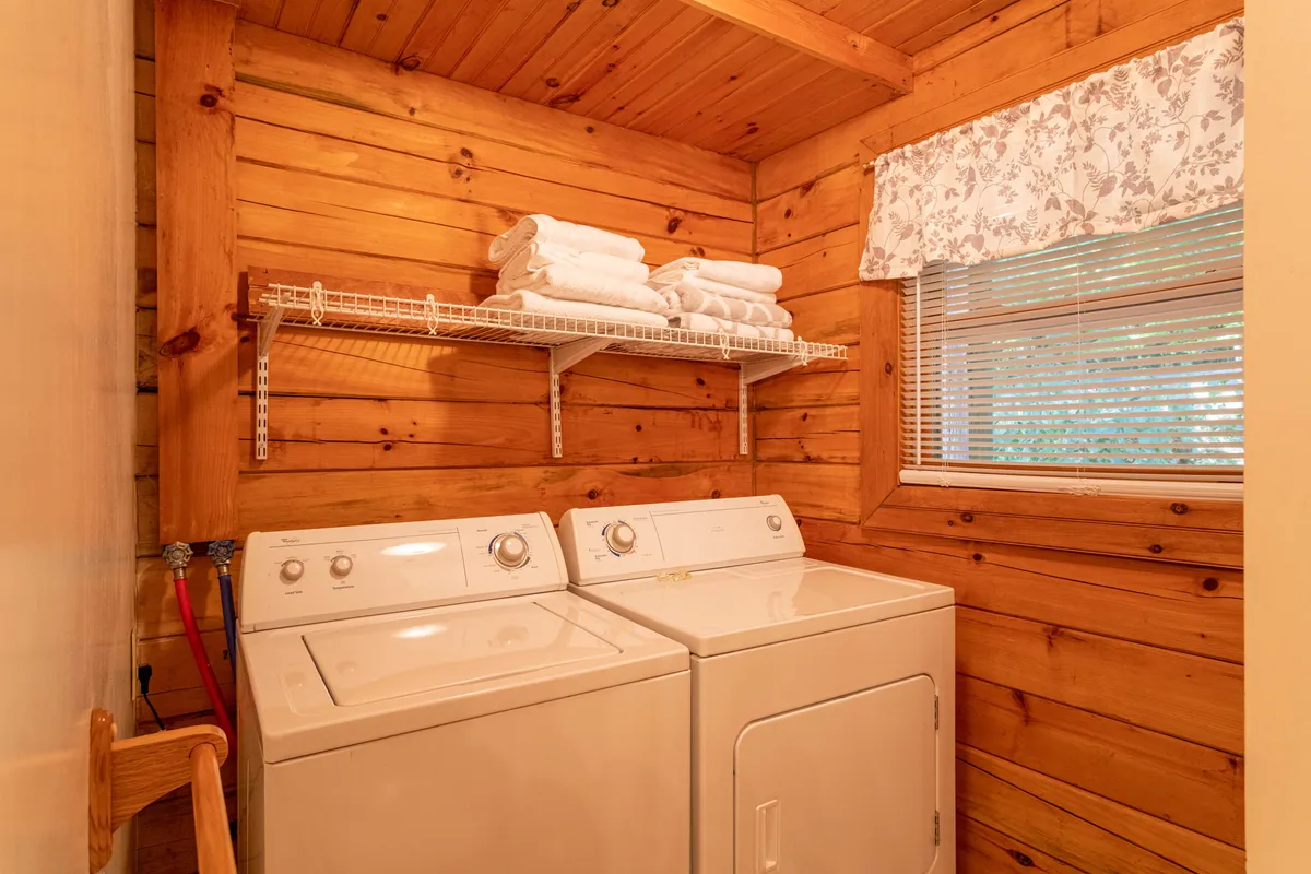 The image features a bright and functional laundry area complete with a modern washing machine, dryer, shelves neatly stocked with towels, and a window adorned with a floral curtain that lets in natural light. The space feels inviting and homey, emphasizing convenience and warmth.