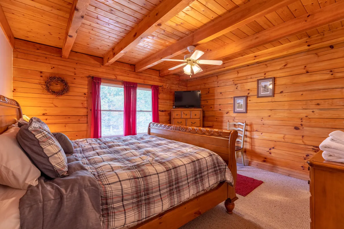 A cozy bedroom with a wooden bed, plaid bedding, expansive windows allowing plenty of natural light, wood-paneled walls, and a ceiling fan. The room feels warm and comfortable, inviting relaxation and restful sleep.