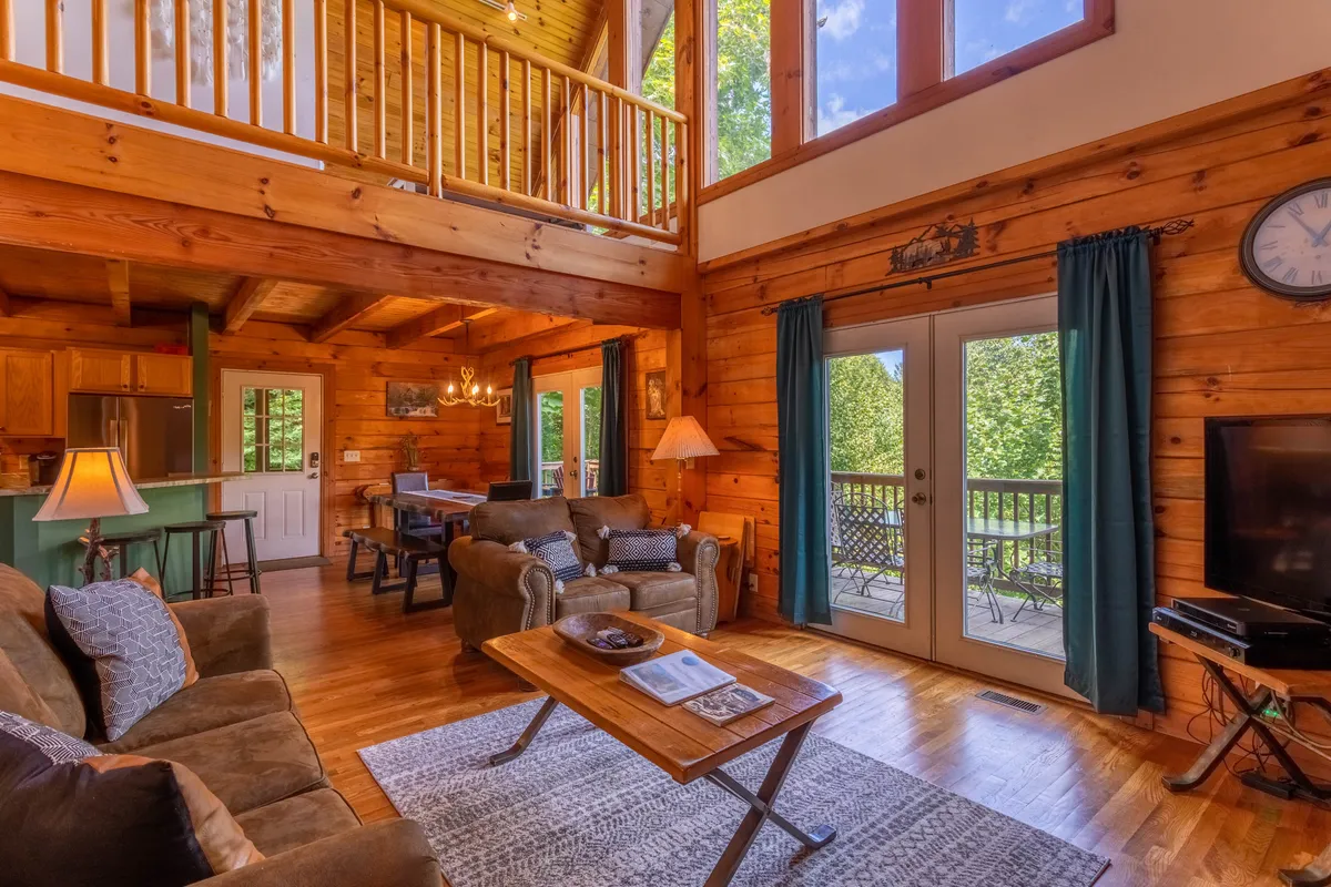 This inviting living area features large floor-to-ceiling windows that flood the space with natural light and frame sweeping mountain views. Wooden beams add rustic charm overhead, while plush seating around a central coffee table creates a cozy gathering spot.