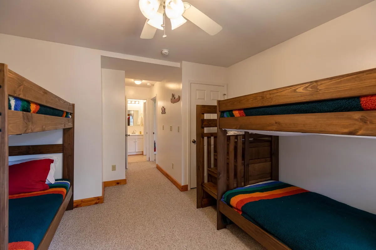 A cozy bunk room with multiple bunk beds dressed in colorful blankets, topped with a ceiling fan above. The space feels playful and welcoming, perfect for kids or teens to share stories, laughter, and memorable nights together.