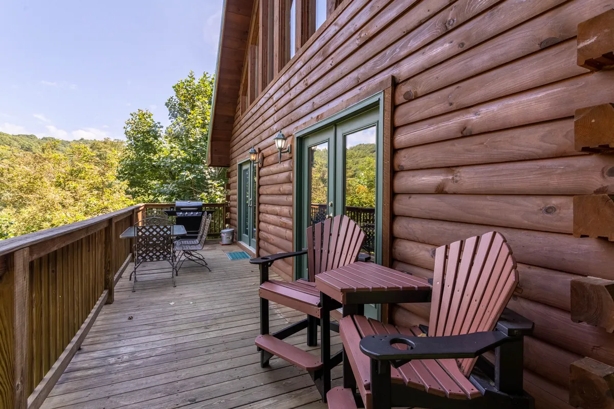 This image showcases a spacious wooden deck attached to the log cabin, furnished with comfortable chairs and an outdoor dining set. The deck is surrounded by lush greenery, inviting guests to relax, socialize, or dine outdoors while taking in tranquil natural views.