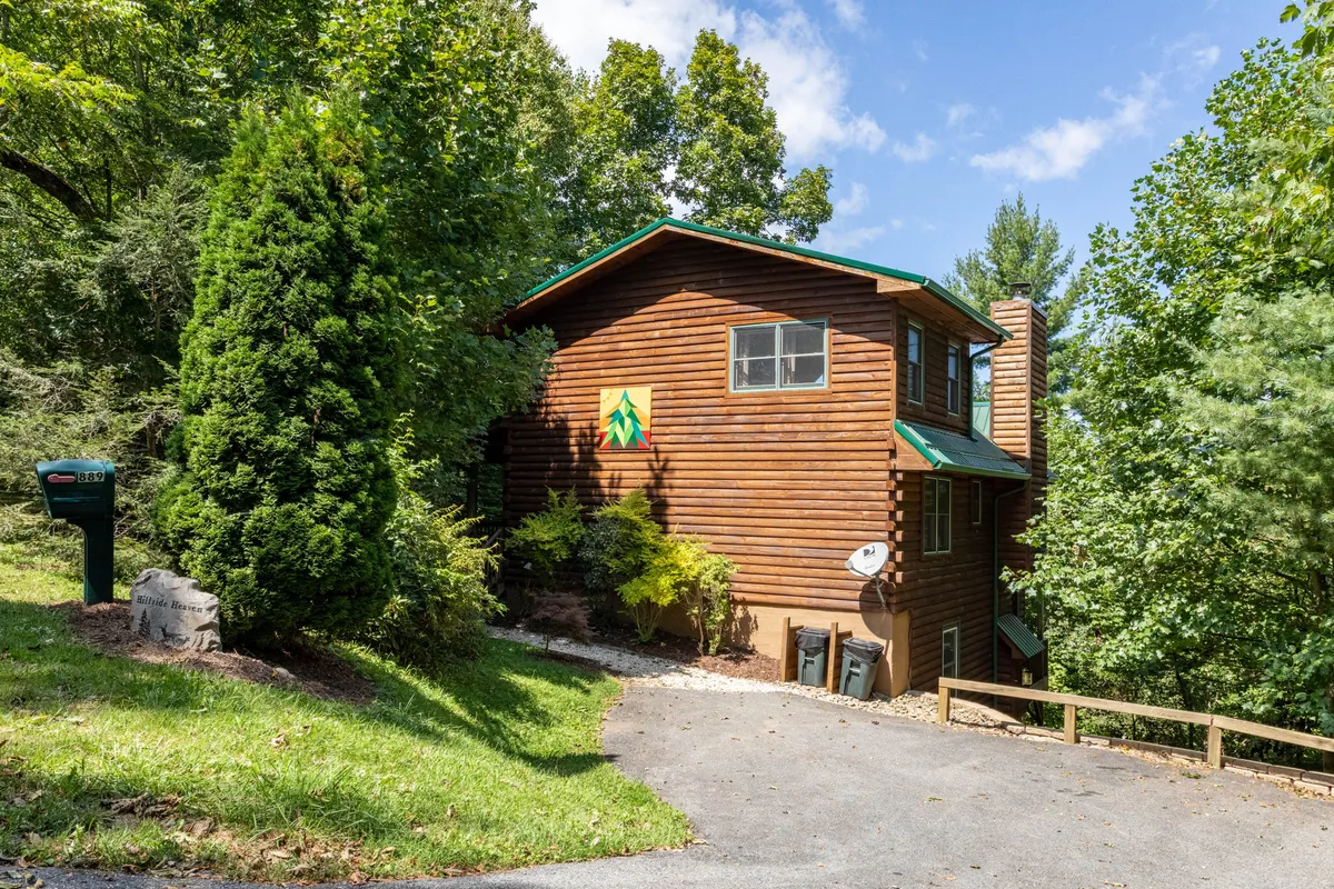 The wooden exterior of the house stands amidst lush greenery, with a green roof and a colorful mountain emblem. The peaceful setting suggests easy access to outdoor spaces and proximity to nature, ideal for launching a day of river adventures.