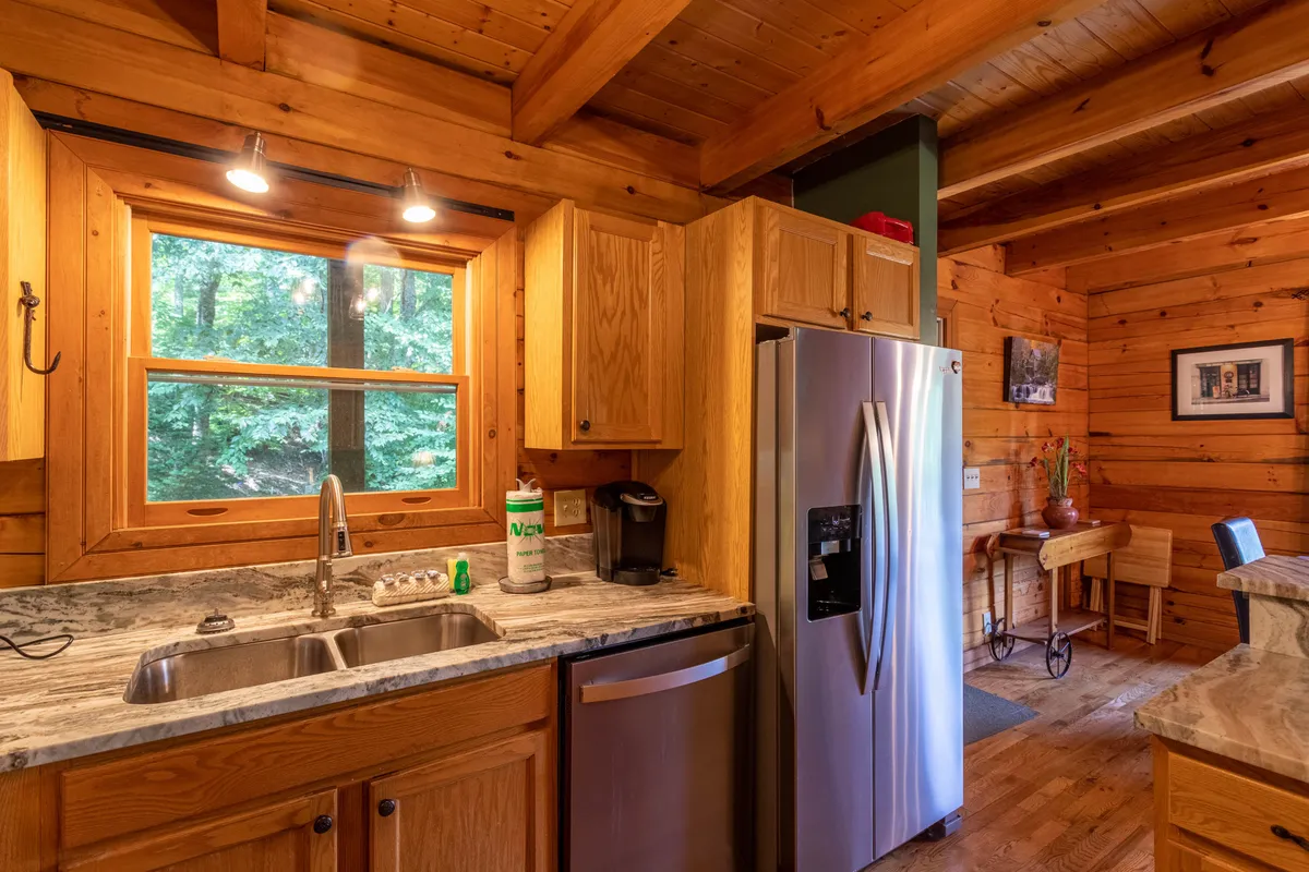 A cozy kitchen area with a sink, refrigerator, and wooden cabinets accented by a window that provides a view of lush trees outside. The natural light emphasizes the inviting, homey feel and suggests easy transition between indoor and outdoor activities.