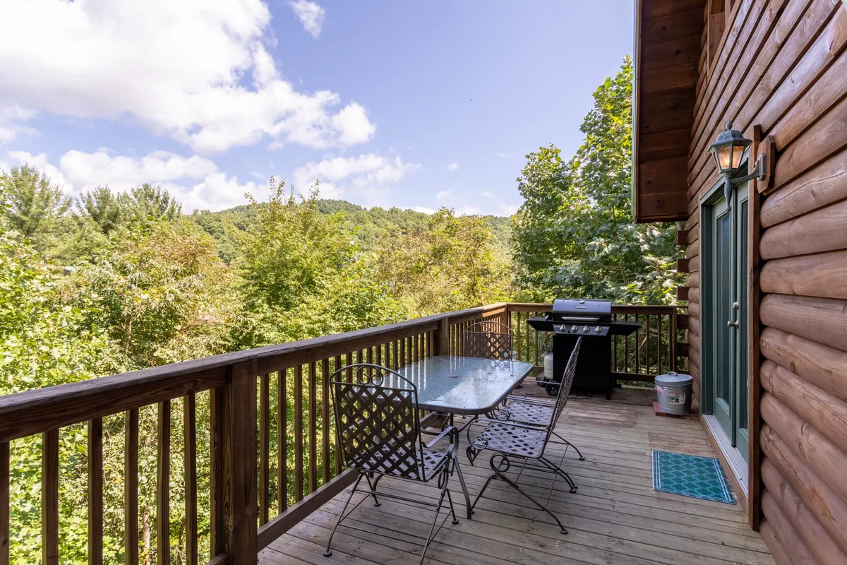 An open-air wooden deck equipped with a table, chairs, and a gas grill overlooks the lush greenery, inviting guests to dine outside or enjoy sunset barbecues.