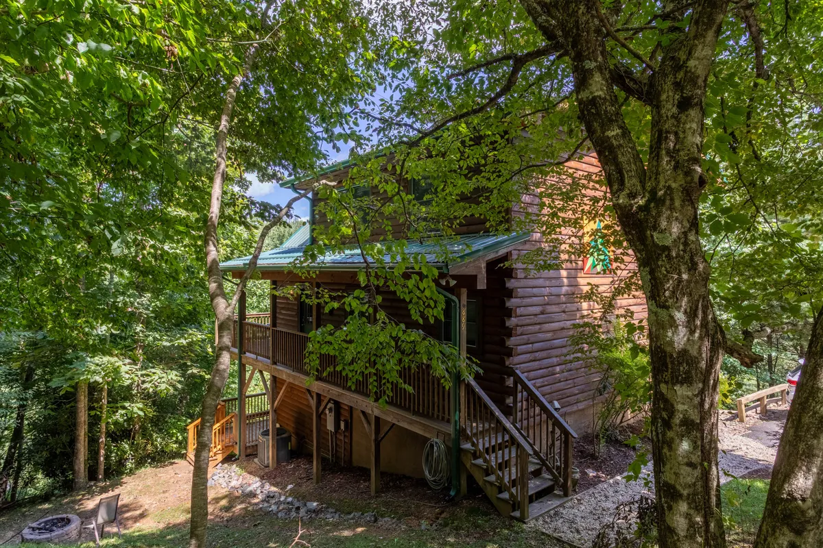 A wide exterior shot of the log cabin with its green metal roof, surrounded by mature trees and a spacious deck area. The setting is serene and exudes the promise of peaceful mornings, stargazing nights, and effortless access to outdoor activities.