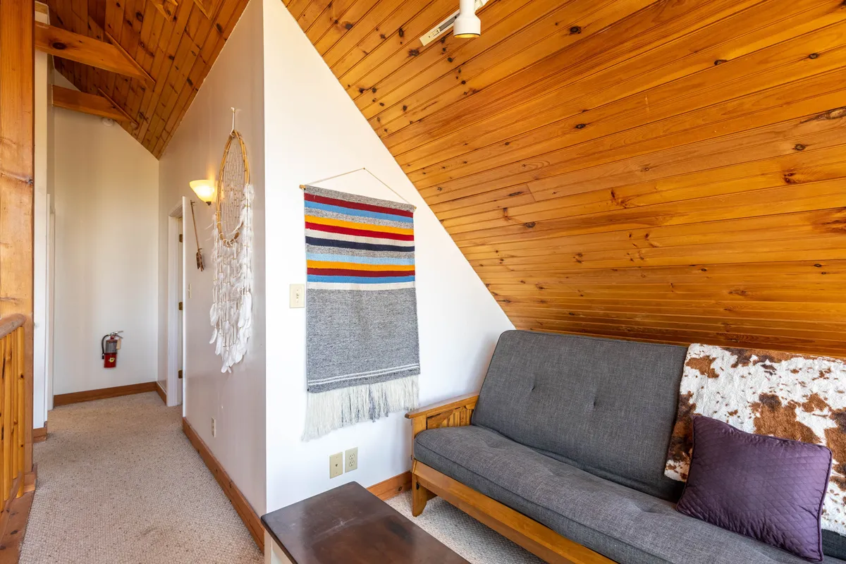 A charming living area featuring a cozy gray sofa, warm wooden ceiling, woven wall art, and carefully chosen decorative accents. The setting is perfect for relaxing, socializing, or snuggling up by the fire, evoking the comforting, intimate atmosphere described in the section.